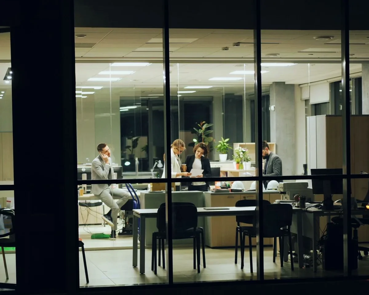 A team talking in a brightly lit office at night, seen through the building's dark windows